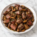 Steak bites with sautéed mushrooms in a glossy garlic-herb sauce, served on a complete white ceramic plate over bright white marble.