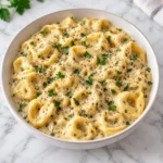 Creamy tortellini sauce with cheese tortellini in a full shallow white ceramic bowl on a bright white marble surface, topped with Parmesan, parsley, herbs, and black pepper, with the complete bowl visible.