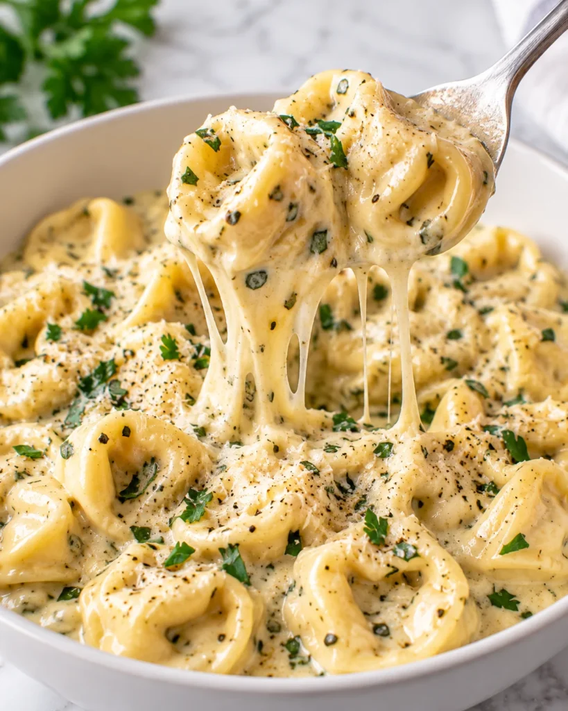 Alt text: Close-up of creamy cheese tortellini being lifted with a silver spoon from a shallow white ceramic bowl on a bright white marble surface, showing glossy sauce, herbs, Parmesan, and soft cheese strands.