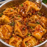 Chili-garlic dumplings in a modern stainless-steel bowl on a bright white marble surface, shown as a complete serving vessel with glossy sauce, chili flakes, sesame-like bits, and scallion garnish.