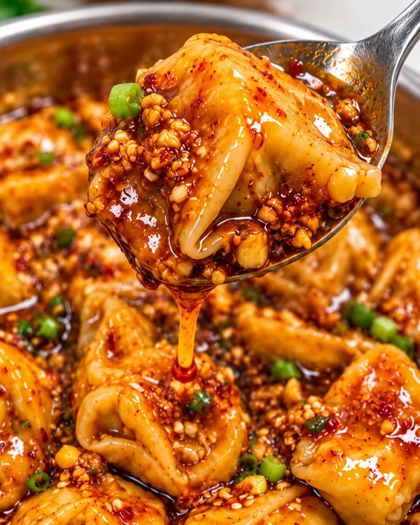 Close-up of a chili-garlic dumpling lifted on a silver spoon from a stainless-steel bowl on a bright white marble surface, with glossy chili oil dripping and sauce-coated dumplings softly visible behind it.