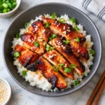 Spicy Korean chicken with glossy caramelized, lightly charred edges served over white rice in a complete dark ceramic bowl, garnished with sesame seeds and green onions on a bright white marble surface.