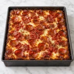 Detroit-style pepperoni pizza baked in a dark rectangular metal pan, fully visible on a bright white marble surface, with bubbly melted cheese, red tomato sauce, crispy cupped pepperoni, and caramelized browned edges.