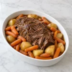 Slow cooker beef pot roast with tender carrots and yellow potatoes in savory juices, served in a white oval dish on white marble.
