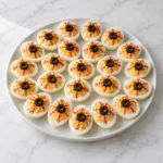 Deviled egg eyeballs with black olive pupils and red veins on a white ceramic platter over white marble.