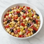 Italian chickpea salad with provolone, salami, olives, peppers, and red onion in a white ceramic bowl on white marble.